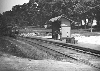 Black and white photo of a small train waiting area along tracks with an approaching steam train.